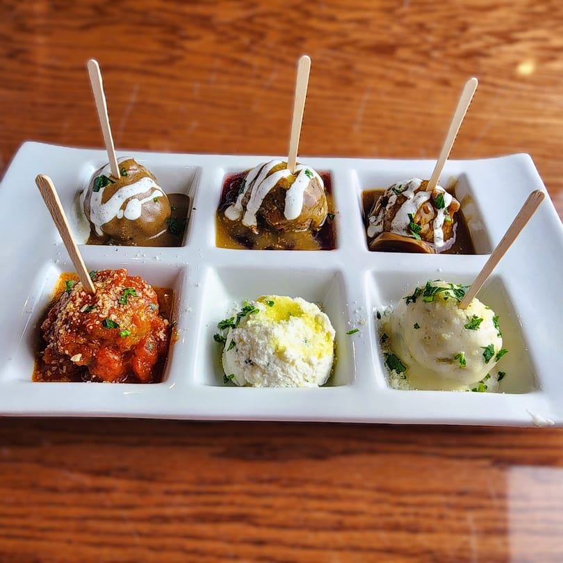 Six Rock N Roll Meatballs, each in a separate square dish, with various sauces and toppings.