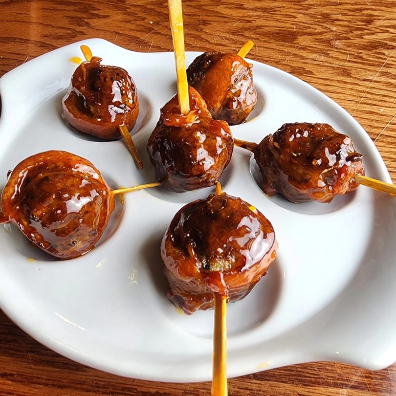 Six glazed meatballs on skewers, served on a white plate, against a wooden table.